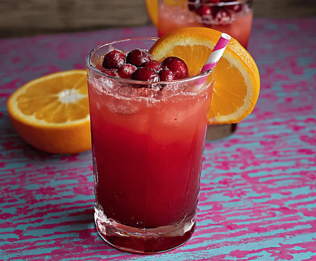 A refreshing glass of cranberry and orange juice garnished with fresh orange slices and cranberries, served in a clear glass pitcher on a bright kitchen countertop.