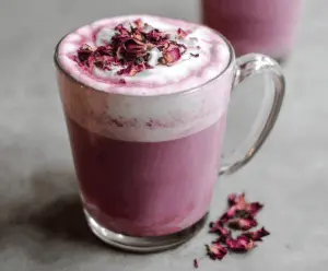 A vibrant Raspberry Hibiscus Latte featuring a pink-hued beverage topped with fresh raspberries and hibiscus flower petals, served in a clear glass mug on a wooden table.